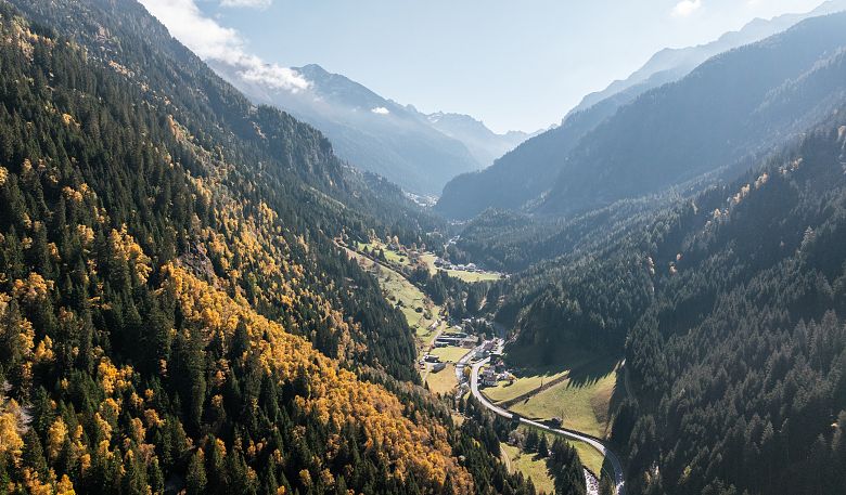 Luftaufnahme eines malerischen Tals in den Alpen, umgeben von grünen und herbstlich gefärbten Wäldern. Eine kleine Ortschaft und Straßen schlängeln sich durch die Landschaft.
