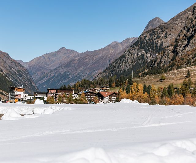 Ein malerisches Bergtal mit einer Langlaufloipe im Vordergrund. Im Hintergrund sind ein Dorf und Wälder in herbstlichen Farben, eingerahmt von hohen Berggipfeln.