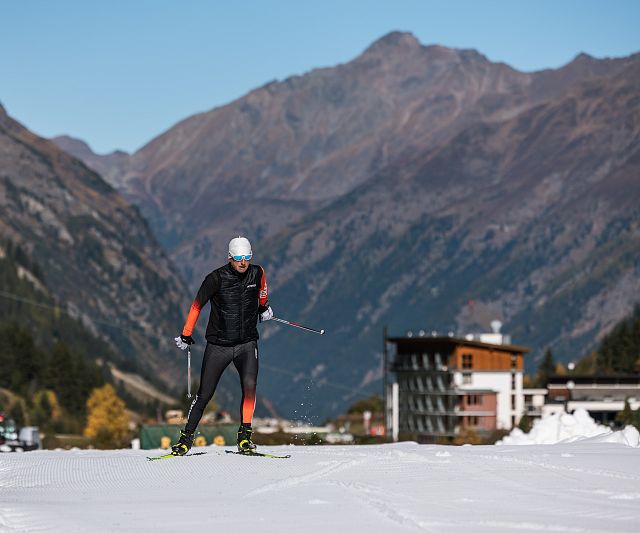 Eine Person in Sportkleidung fährt auf Skiern eine schneebedeckte Loipe hinunter, umgeben von bergiger Landschaft und einigen Gebäuden im Hintergrund bei klarem Himmel.