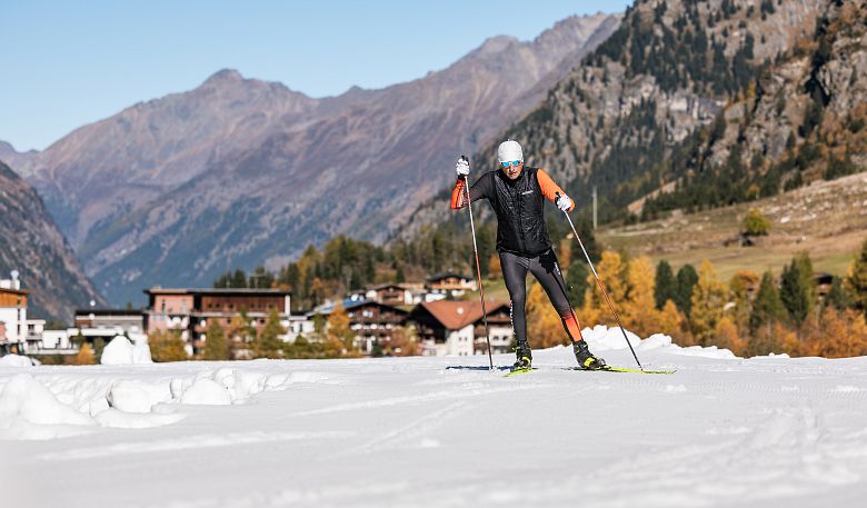 Ein Langläufer in Wintersportkleidung fährt über eine schneebedeckte Landschaft. Im Hintergrund sind Herbstbäume, ein Dorf und Berge unter klarem Himmel zu sehen.