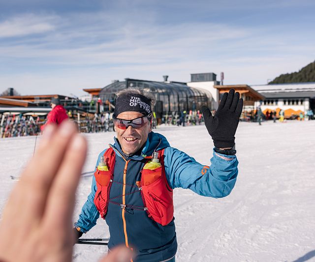 Ein Mensch in Winterkleidung steht auf einer Skipiste und lächelt. Im Hintergrund sind Gebäude und Bäume zu sehen. Eine Hand im Vordergrund deutet ein High Five an.