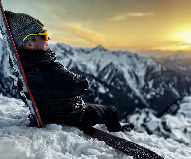 Person auf einem Berg lehnt mit Snowboard und schaut in den Sonnenuntergang über schneebedeckte Gipfel. Ruhige Winterlandschaft im Abendlicht.