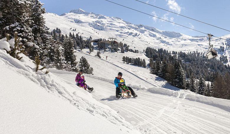 Zwei Personen mit Kindern rodeln unter einem klaren blauen Himmel die schneebedeckte Rodelbahn am Hochzeiger im Pitztal hinunter.