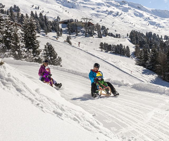 Zwei Personen mit Kindern rodeln unter einem klaren blauen Himmel die schneebedeckte Rodelbahn am Hochzeiger im Pitztal hinunter.