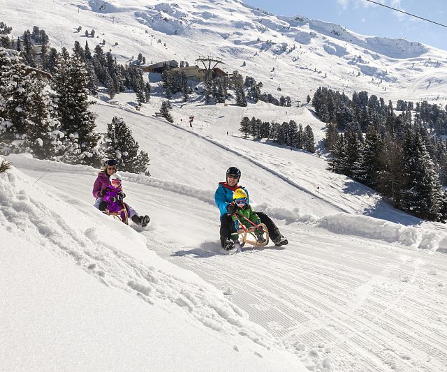 Zwei Personen mit Kindern rodeln unter einem klaren blauen Himmel die schneebedeckte Rodelbahn am Hochzeiger im Pitztal hinunter.