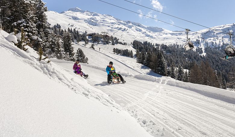 Zwei Personen mit Kindern rodeln unter einem klaren blauen Himmel die schneebedeckte Rodelbahn am Hochzeiger im Pitztal hinunter.