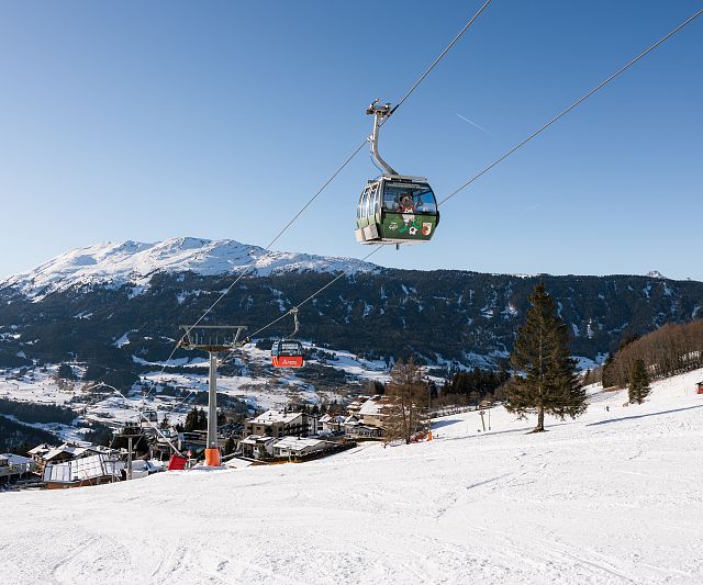 Eine Gondelbahn schwebt über einer schneebedeckten Skipiste. Im Hintergrund sind verschneite Berge und ein klarer blauer Himmel zu sehen. Einige Bäume säumen die Piste.