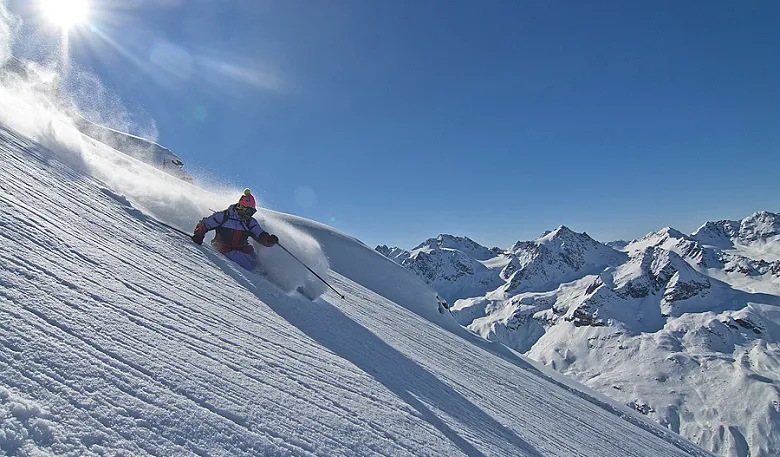 Ein Skifahrer fährt eine verschneite Bergpiste hinunter, umgeben von schneebedeckten Gipfeln unter strahlend blauem Himmel.