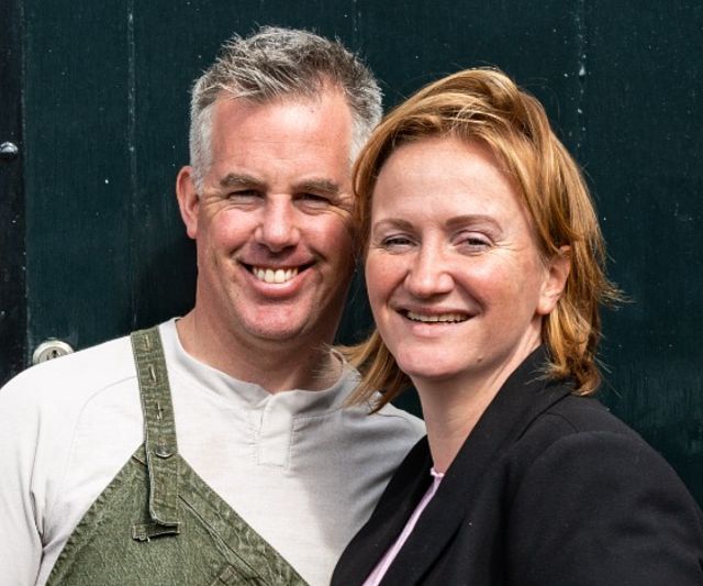 Es zeigt einen Arjan Wennekes in einem grünen Overall und Laura Wennekes in einem schwarzen Blazer, beide lächelnd. Die beiden sind die Besitzer vom Landhaus Edelweiß in Tieflehn.