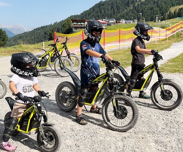 Mehrere Kinder mit Helmen stehen auf einem Kiesweg vor einer bergigen Landschaft. Sie sitzen auf gelben Elektromotorrädern und tragen Schutzkleidung.