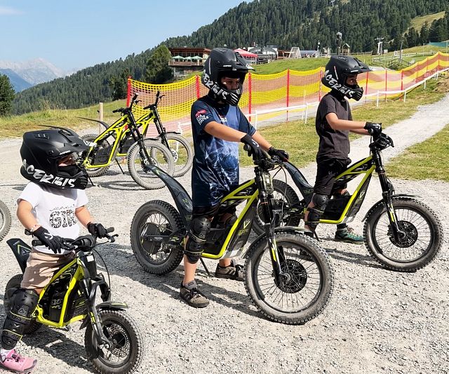 Mehrere Kinder mit Helmen stehen auf einem Kiesweg vor einer bergigen Landschaft. Sie sitzen auf gelben Elektromotorrädern und tragen Schutzkleidung.