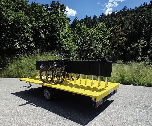Ein gelber Anhänger trägt mehrere angeschlossene Fahrräder auf einer ländlichen Straße vor einem Hintergrund mit üppig grünen Bäumen und blauem Himmel.