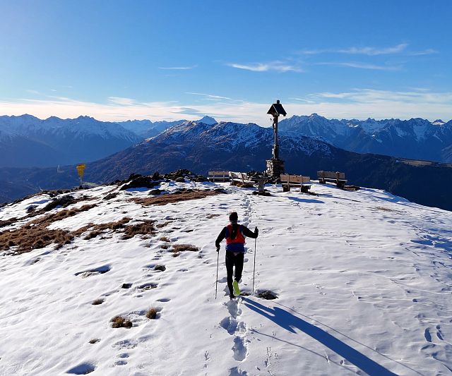 A trail runner walks with poles on a snowy mountain peak to a summit cross. Wide mountain ranges are visible in the background under a clear blue sky.