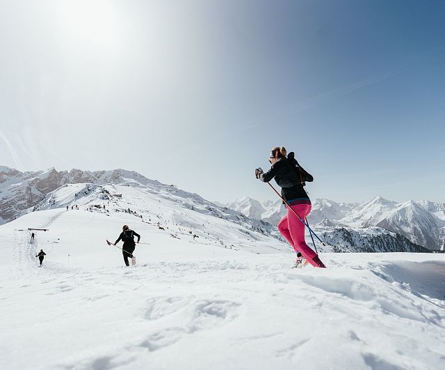Menschen laufen mit Skistöcken durch eine schneebedeckte Berglandschaft bei klarem Himmel. Im Hintergrund sind beeindruckende Gipfel zu sehen.