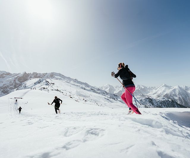 Menschen laufen mit Skistöcken durch eine schneebedeckte Berglandschaft bei klarem Himmel. Im Hintergrund sind beeindruckende Gipfel zu sehen.