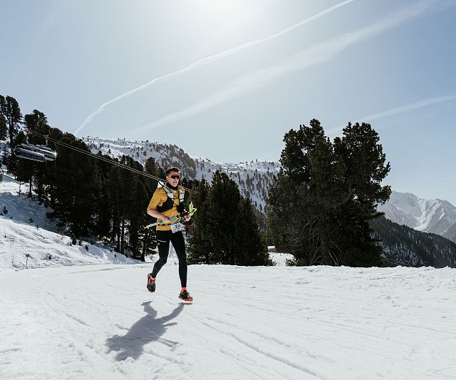 Person läuft in winterlicher Landschaft auf einem schneebedeckten Bergpfad, umgeben von Bäumen und Bergen, bei sonnigem Wetter.