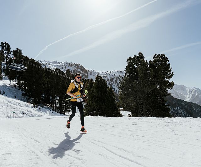 Person läuft in winterlicher Landschaft auf einem schneebedeckten Bergpfad, umgeben von Bäumen und Bergen, bei sonnigem Wetter.