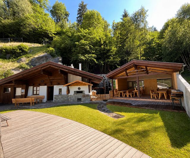 Two rustic wooden huts stand in the middle of a green garden, surrounded by trees. A terrace with wooden tables provides seating and exudes a sense of tranquillity.