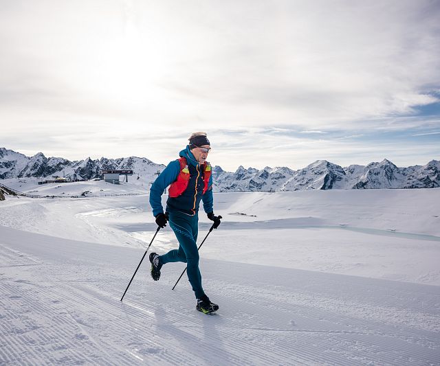 Eine Trailrunner im blauen Anzug läuft mit Skistöcken über schneebedeckte Berge. Der Himmel ist bewölkt, und im Hintergrund sind beeindruckende Berggipfel zu sehen.