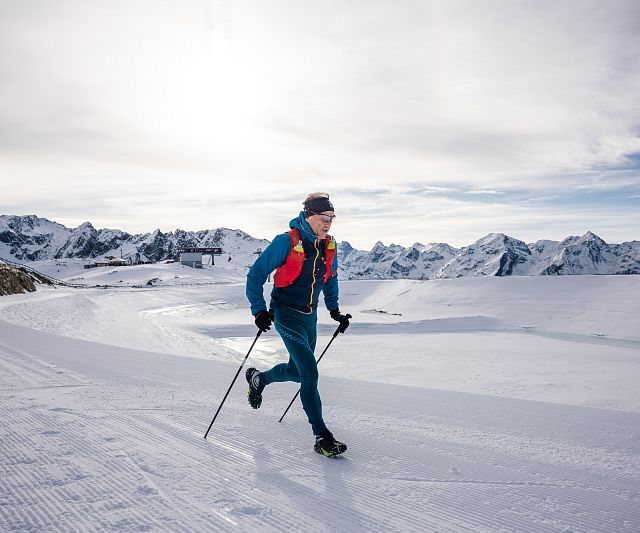 Eine Trailrunner im blauen Anzug läuft mit Skistöcken über schneebedeckte Berge. Der Himmel ist bewölkt, und im Hintergrund sind beeindruckende Berggipfel zu sehen.