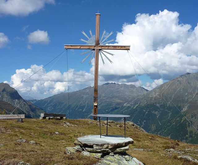 Ein Holz-Kreuz steht auf einer Anhöhe. Es ist umgeben von Bergen und Wiesen unter einem blauen Himmel mit Wolken. Der Ort strahlt eine ruhige, alpine Atmosphäre aus.