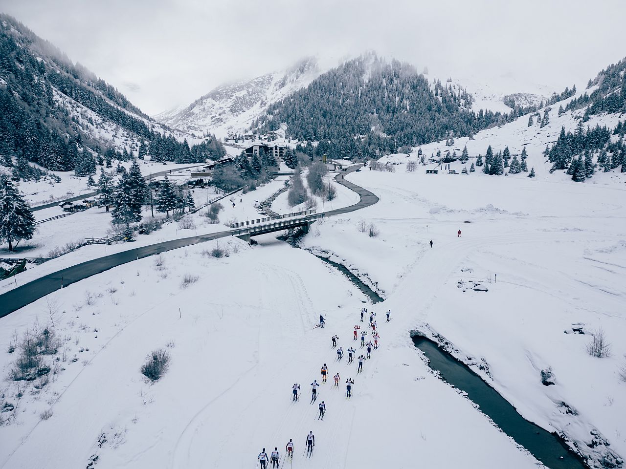 Eine Gruppe von Menschen skatet auf verschneiten Wegen durch eine winterliche Berglandschaft. Im Hintergrund schlängelt sich ein Fluss entlang schneebedeckter Bäume.