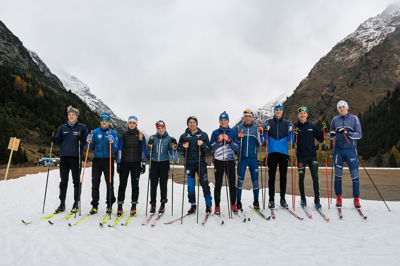 A group of eleven cross-country skiers stand next to each other on a snow-covered surface. They are wearing winter clothing and skis, with mountains and clouds in the background.