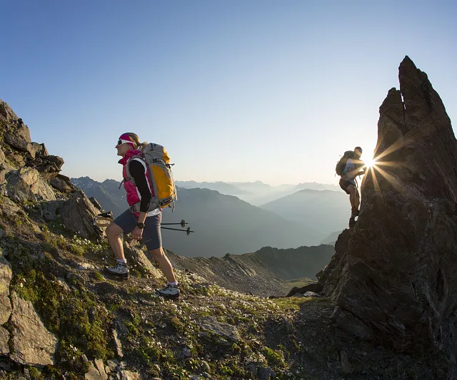 Zwei Wanderer erklimmen einen Bergpfad in den Alpen. Die Sonne geht auf und strahlt durch einen Felsen. Ein Naturpanorama mit Bergen und Tälern im Hintergrund.