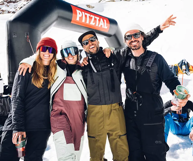 Vier Personen in Winterkleidung posieren lächelnd im Schnee vor einem Torbogen mit 'Pitztal'-Schriftzug. Sie wirken fröhlich und entspannt.