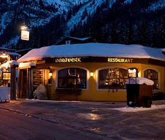 Ein gelb erleuchtetes Restaurant mit schneebedecktem Dach in einem bergigen Winterdorf. Umgeben schneebedeckten Bäumen und einer ruhigen, nächtlichen Atmosphäre.