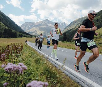 Eine Gruppe von Läufern joggt auf einer Straße durch ein malerisches Tal, umgeben von Bergen. Am Bildrand steht: "20 Jahre Pitztal Gletscher Marathon".