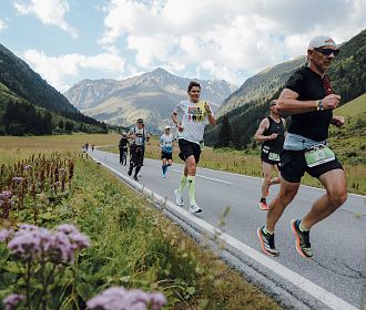 Eine Gruppe von Läufern joggt auf einer Straße durch ein malerisches Tal, umgeben von Bergen. Am Bildrand steht: "20 Jahre Pitztal Gletscher Marathon".
