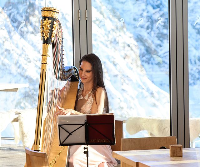 Eine Frau spielt Harfe in einem sonnigen Raum mit Panoramafenstern und verschneiten Bergen im Hintergrund. Auf dem Tisch vor ihr stehen eine Wasserflasche und ein Notenständer.