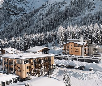 Eine verschneite Berglandschaft mit mehreren modernen Holzgebäuden, umgeben von schneebedeckten Bäumen und majestätischen Bergen im Hintergrund. Winter Ambiente.