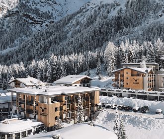 Eine verschneite Berglandschaft mit mehreren modernen Holzgebäuden, umgeben von schneebedeckten Bäumen und majestätischen Bergen im Hintergrund. Winter Ambiente.