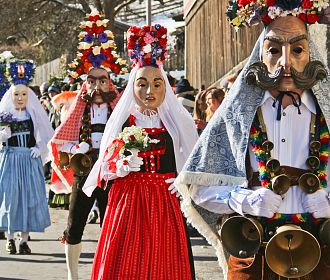 Eine Gruppe von Menschen in traditionellen Kostümen und Masken bei einem kulturellen Festumzug. Bunte Kleidung und große Hüte mit Blumen schmücken die Teilnehmer.