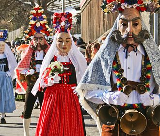 Eine Gruppe von Menschen in traditionellen Kostümen und Masken bei einem kulturellen Festumzug. Bunte Kleidung und große Hüte mit Blumen schmücken die Teilnehmer.