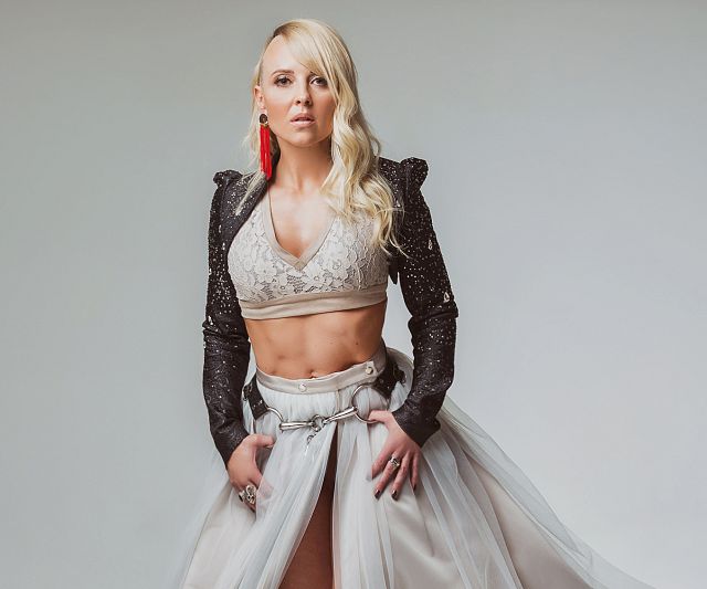 Eine Frau in einem eleganten Outfit posiert in einem Fotostudio. Sie trägt ein langes, fließendes Kleid mit einem schwarzen Oberteil. Der Hintergrund ist schlicht grau.