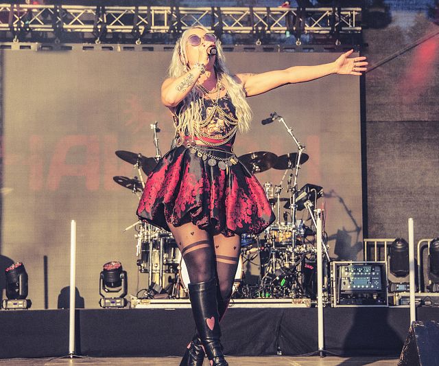 Sängerin auf einer Bühne in auffälligem Outfit mit rotem Rock und schwarzen Stiefeln. Sie singt in ein Mikrofon, während eine Band ihre Performance begleitet. Bühne beleuchtet.