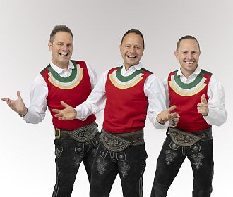 Drei Männer in traditionellen Trachten mit roten Westen und weißen Hemden posieren fröhlich vor einem neutralen Hintergrund. Sie tragen schwarze Lederhosen und haben ihre Daumen hoch.