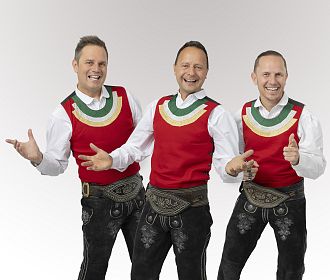 Drei Männer in traditionellen Trachten mit roten Westen und weißen Hemden posieren fröhlich vor einem neutralen Hintergrund. Sie tragen schwarze Lederhosen und haben ihre Daumen hoch.