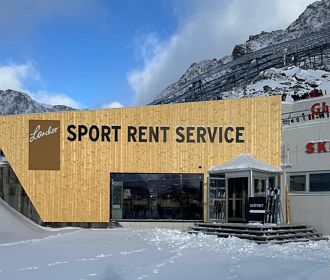 Modernes Holzgebäude mit der Aufschrift "Sport Rent Service". Vor dem Eingang steht ein überdachter Bereich mit Skiern. Schnee bedeckt den Boden, Berge im Hintergrund.