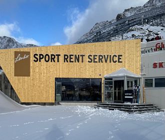 Modernes Holzgebäude mit der Aufschrift &quot;Sport Rent Service&quot;. Vor dem Eingang steht ein überdachter Bereich mit Skiern. Schnee bedeckt den Boden, Berge im Hintergrund.