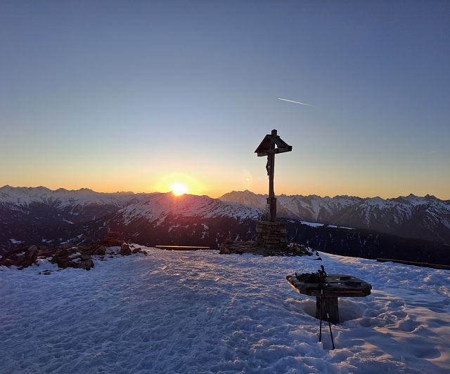 Verschneiter Berggipfel bei Sonnenuntergang mit einem Gipfelkreuz im Vordergrund. untergehende taucht den Himmel die Berge amont in warme.