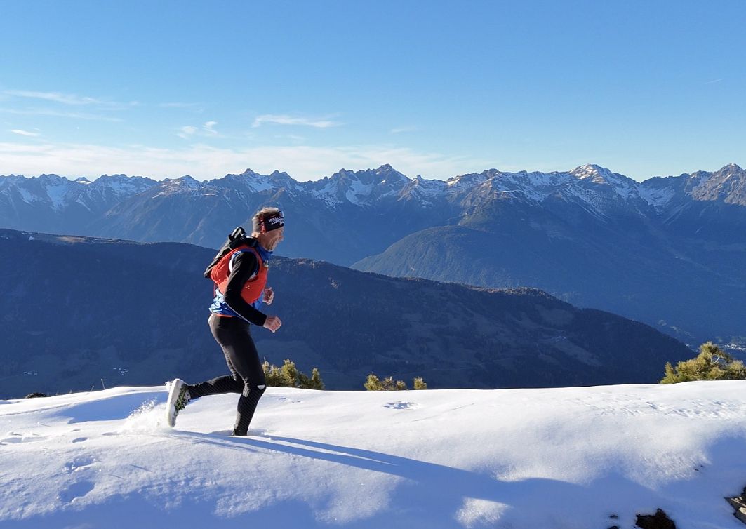 Ein Läufer in Sportkleidung joggt über eine schneebedeckte, sonnige Berglandschaft mit klar blauem Himmel im Hintergrund. Die Alpen erstrecken sich in der Ferne.