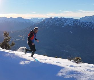 Ein Trail Runner läuft in warmer Kleidung über einen schneebedeckten Berg. Die Sonne scheint am klaren Himmel, mit einer beeindruckenden Berglandschaft im Hintergrund.