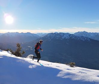 Ein Trail Runner läuft in warmer Kleidung über einen schneebedeckten Berg. Die Sonne scheint am klaren Himmel, mit einer beeindruckenden Berglandschaft im Hintergrund.