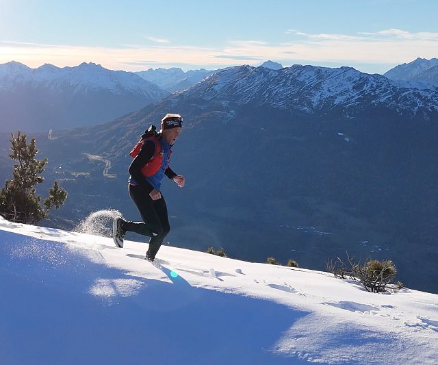 Ein Trail Runner läuft in warmer Kleidung über einen schneebedeckten Berg. Die Sonne scheint am klaren Himmel, mit einer beeindruckenden Berglandschaft im Hintergrund.