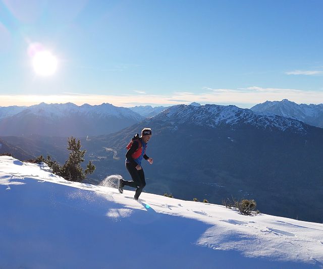 Ein Trail Runner läuft in warmer Kleidung über einen schneebedeckten Berg. Die Sonne scheint am klaren Himmel, mit einer beeindruckenden Berglandschaft im Hintergrund.