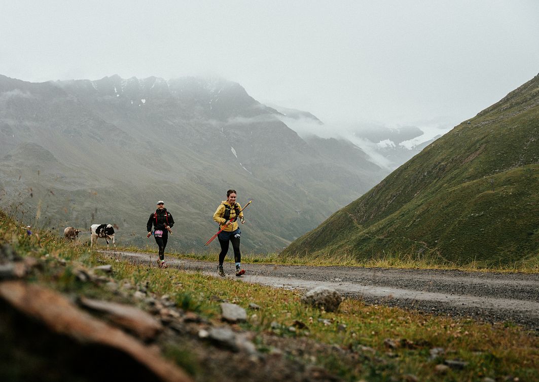 Zwei Trailrunner laufen auf einem nebligen Bergpfad, umgeben von grünen Hügeln und bewölktem Himmel.